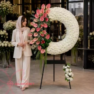 خانمی با کت‌وشلوار و شال، کنار تاج گل کد 002 در گلفروشی گلدون ایستاده و به خوشه‌ی آنتریوم صورتی و گل‌های داوودی سفید تاج نگاه می‌کند تا اندازه و حجم واقعی محصول نمایش داده شود