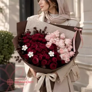 دسته گل رز ژولیت کد 014 از فروشگاه گلدون؛ ترکیبی پرحجم از گل رز ژولیت قرمز شرابی و گلهای صورتی روشن در کنار برگهای تیرهی تزئینی، در بستهبندی کاغذ قهوهای شکلاتی با روبان ساتن در دستان مدل خانم