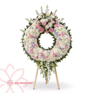 تاج گل کد 009 از فروشگاه گلدون؛ تاج گل حلقه‌ای با داوودی سفید، رز صورتی و کرمی و گل‌های فصلی روی پایه چوبی سه‌پایه؛ رنگ‌بندی پاستلی و آرام